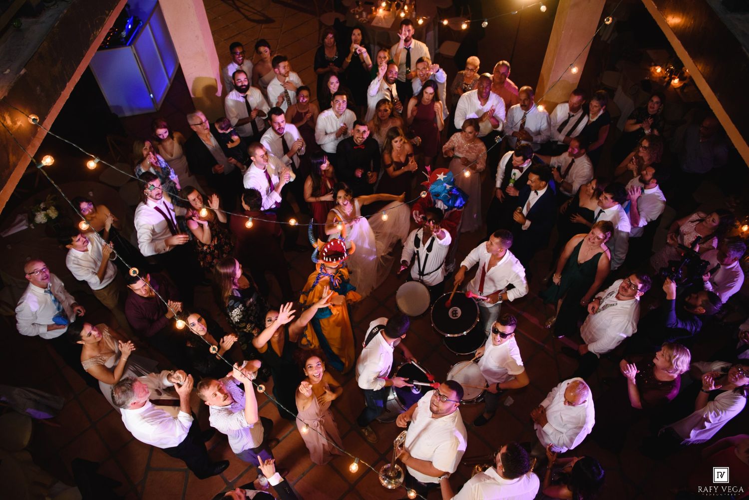 Overhead shot of a crowded wedding reception with guests mingling and dancing under warm string lights.