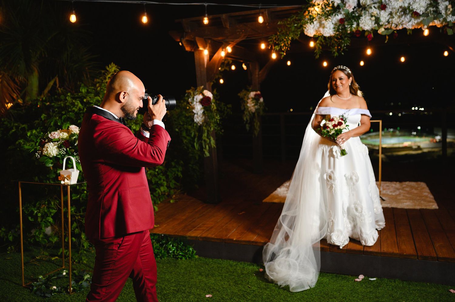 A photographer in a red suit captures a bride in a white gown at an outdoor evening wedding reception.