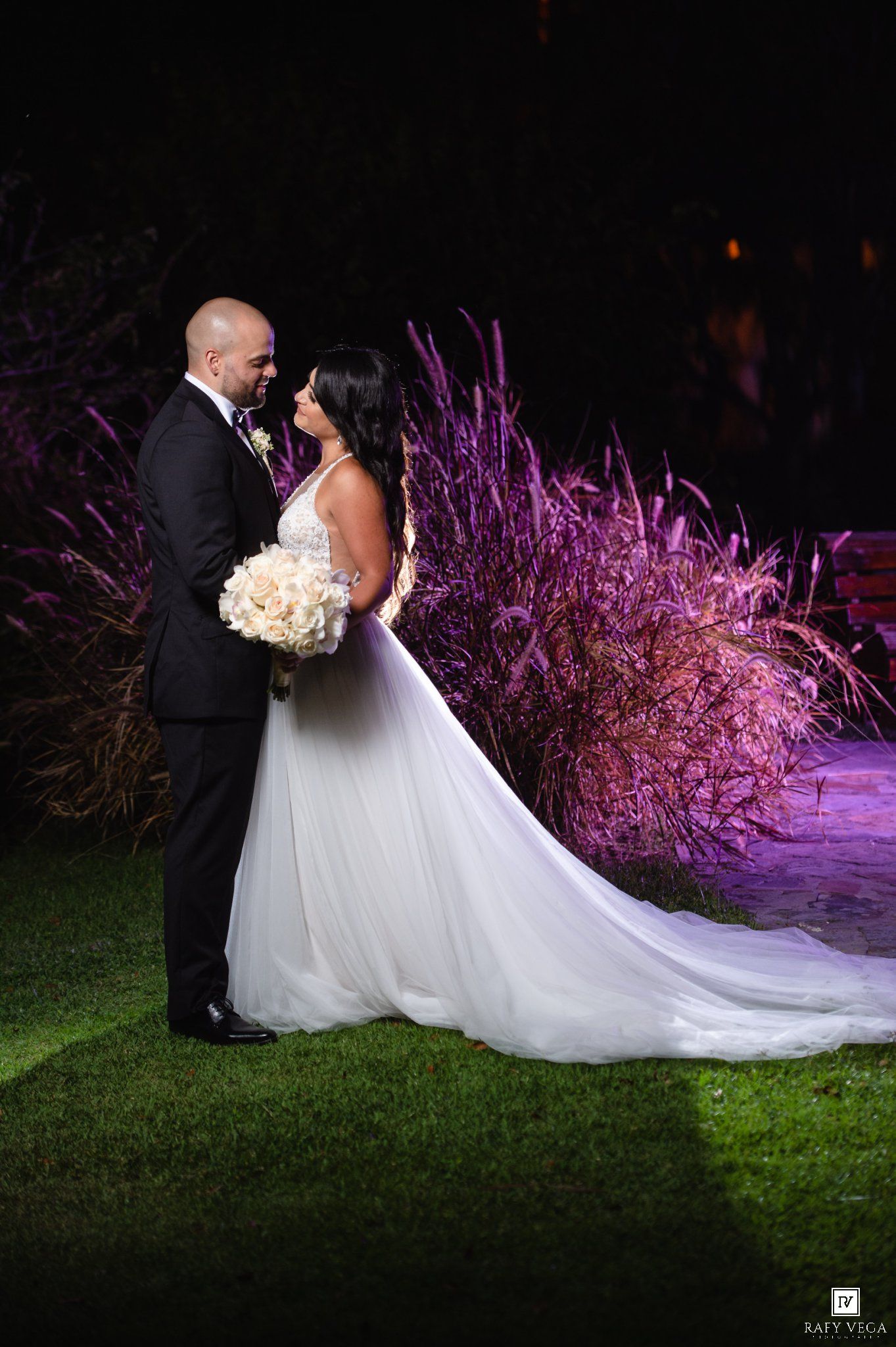 Bride and groom share a romantic moment at night, surrounded by purple-lit ornamental grasses on a lush green lawn.