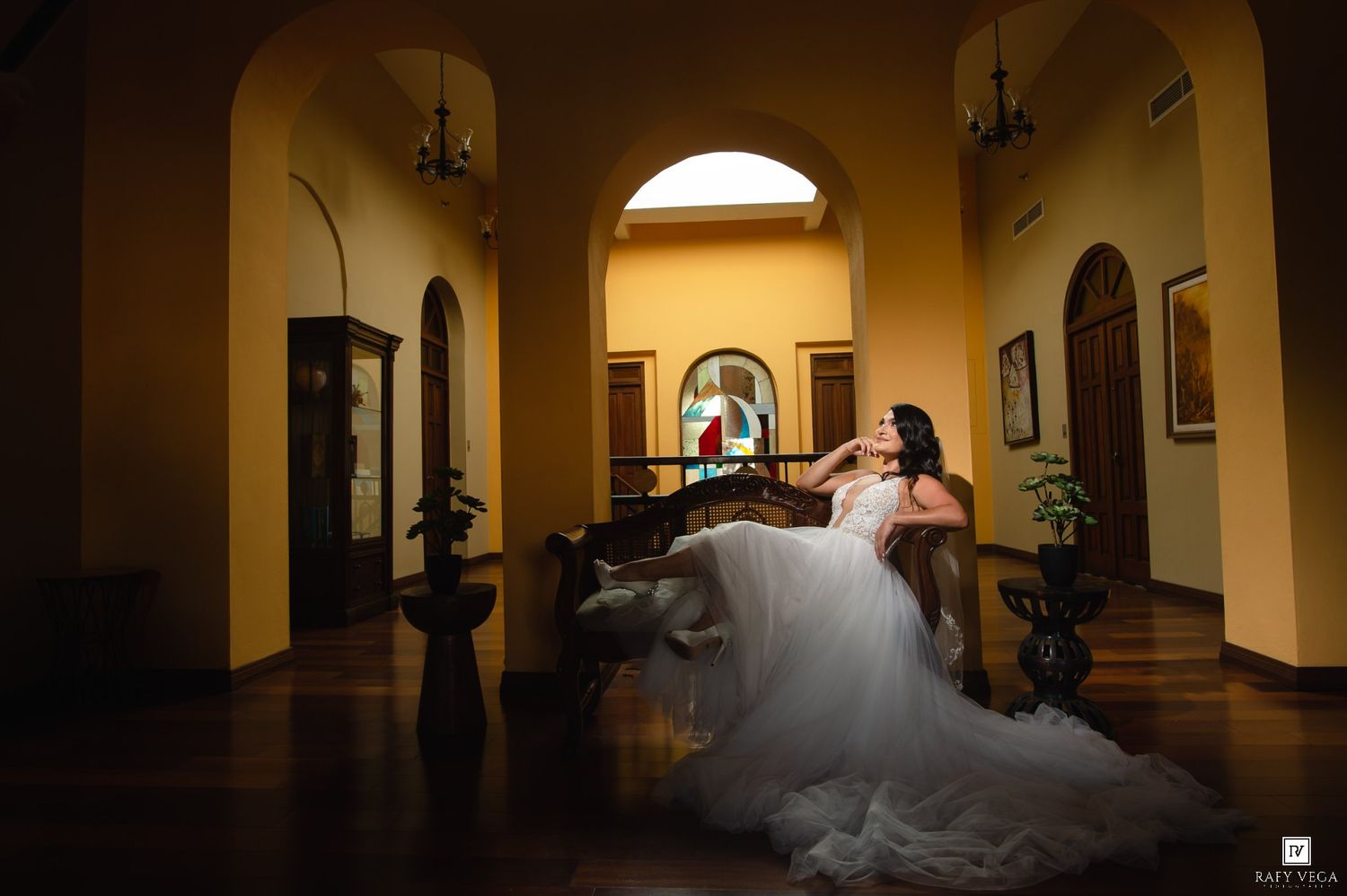 Bride in white gown seated elegantly in a warmly lit colonial hallway with arched windows and wooden decor.