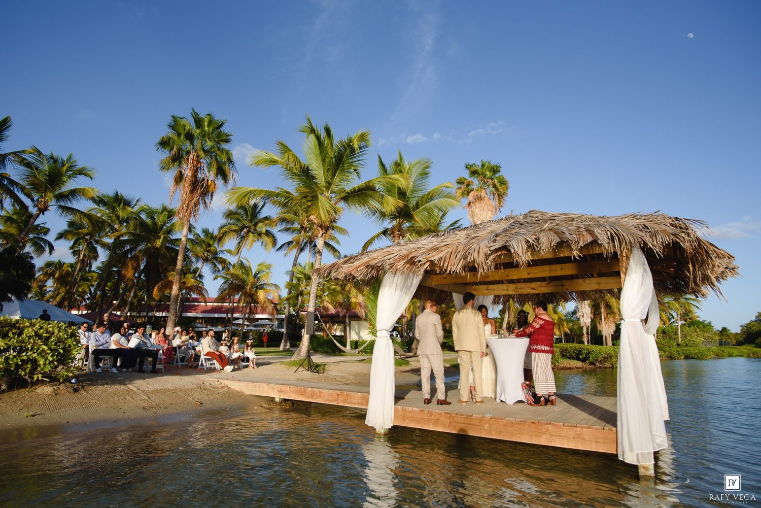 Outdoor waterfront wedding ceremony under a thatched gazebo on a dock, surrounded by palm trees and blue skies.