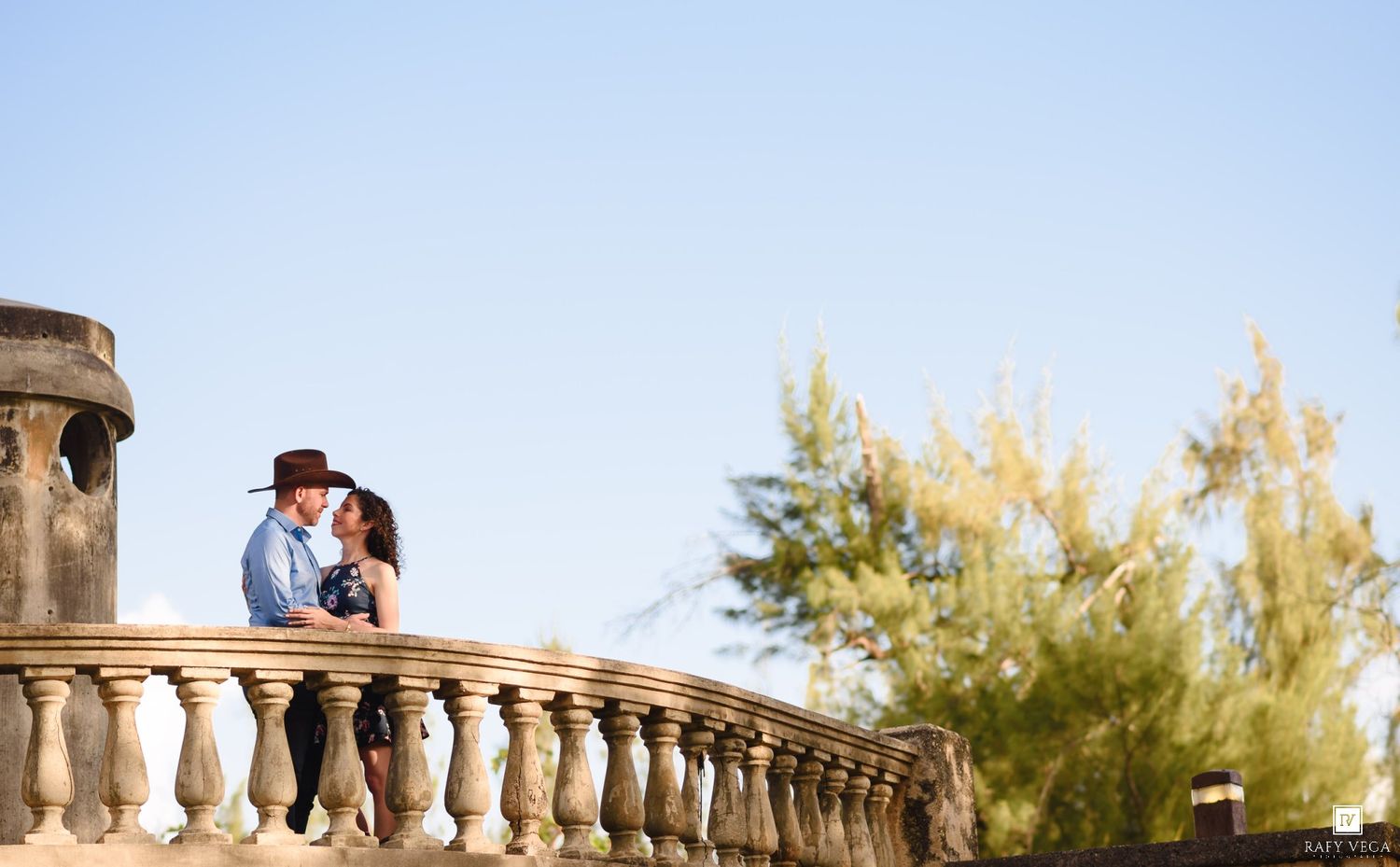 Couple sharing a romantic moment on an ornate stone balustrade balcony surrounded by lush greenery and clear blue sky.
