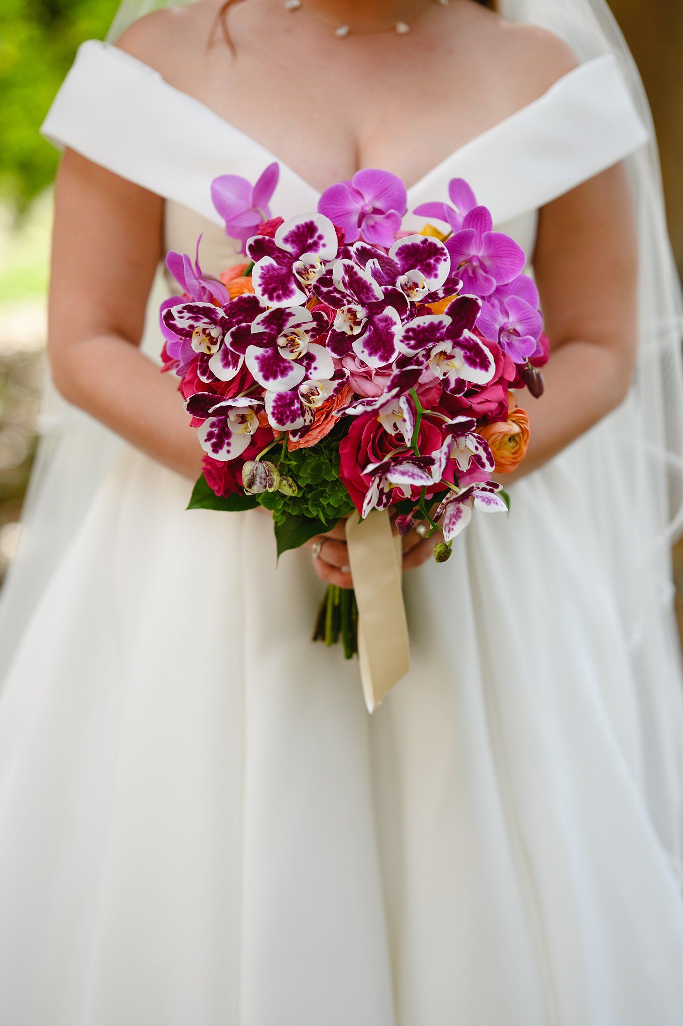 Bride holding vibrant purple and white orchid wedding bouquet with orange accents against white off-shoulder gown.