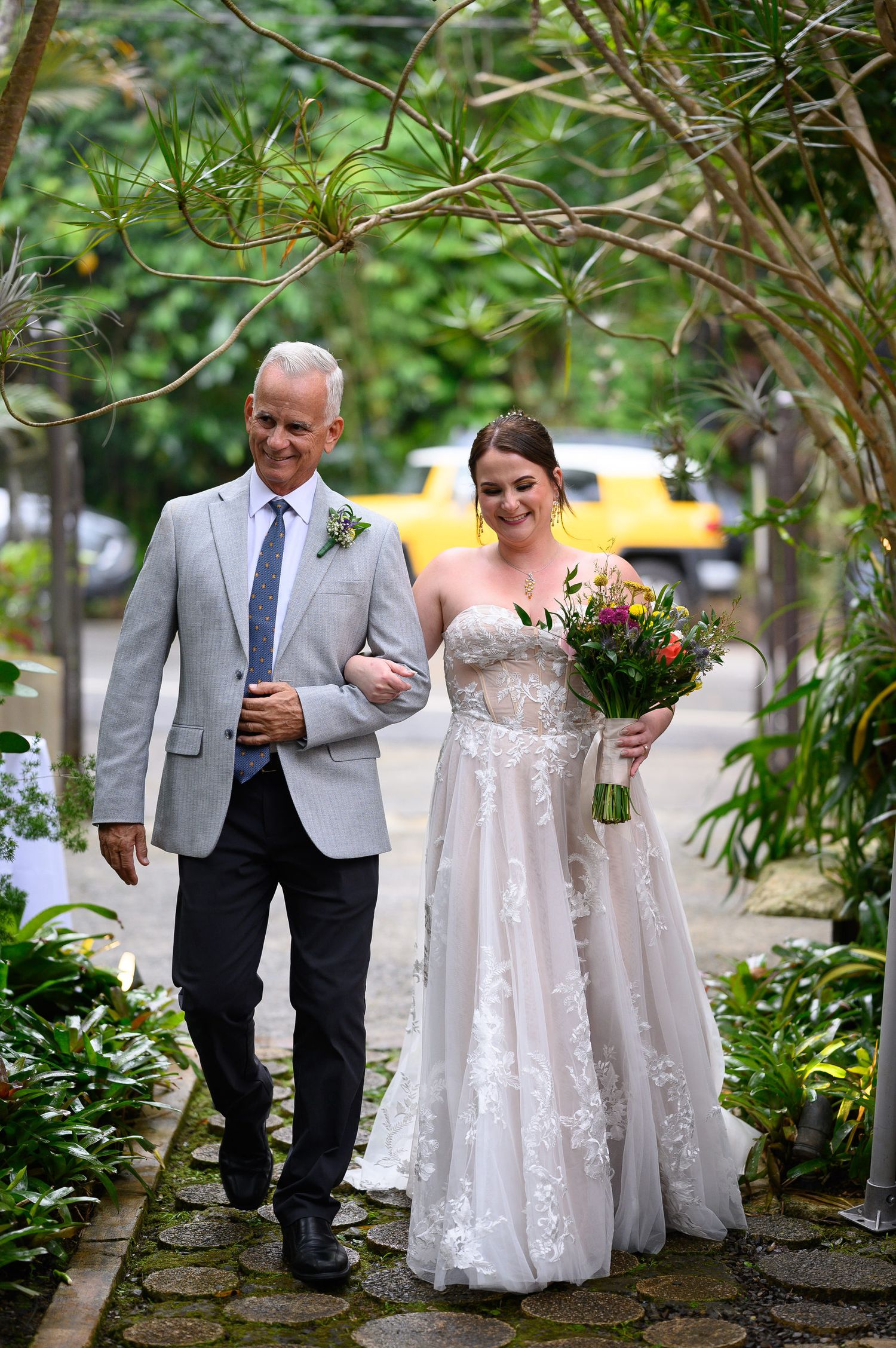 Bride in lace gown holding colorful bouquet walks arm-in-arm with suited man along a lush garden pathway.