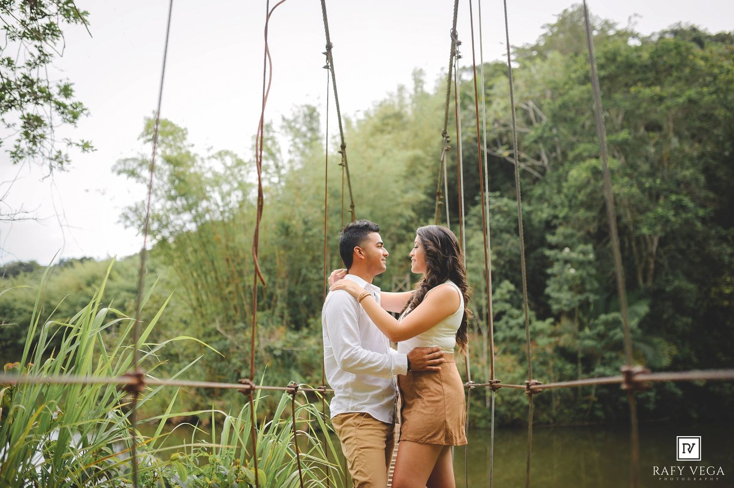 Couple embracing near a tranquil pond surrounded by lush green bamboo trees during an outdoor engagement photoshoot.
