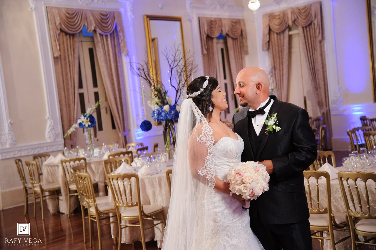 Bride and groom share a romantic moment in an elegant wedding reception hall with blue floral arrangements.
