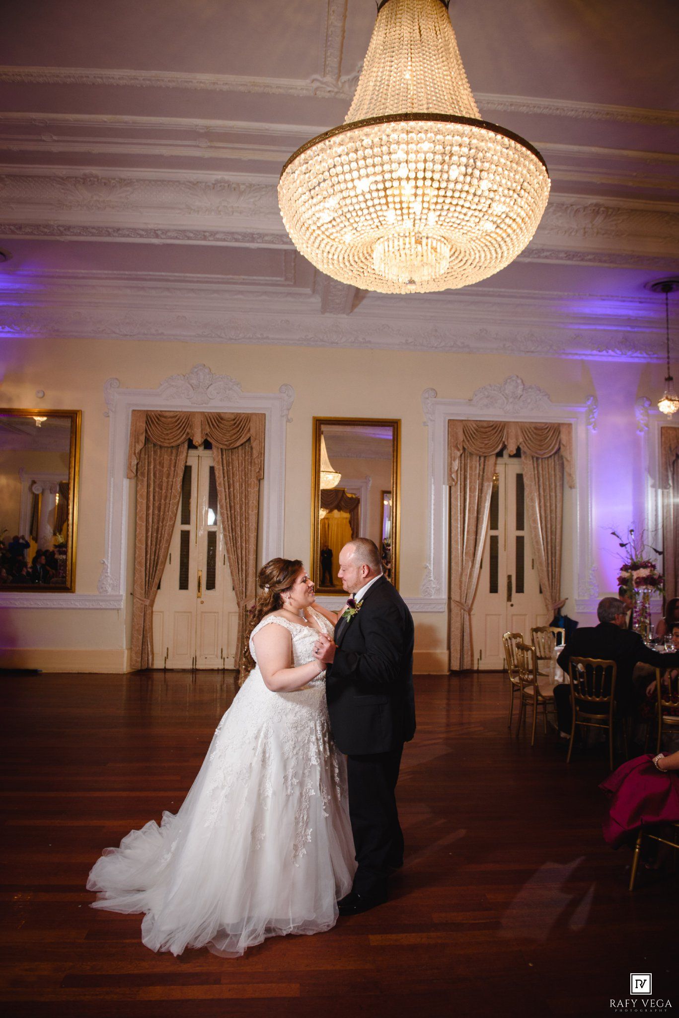 Bride and groom share first dance under elegant crystal chandelier in ornate ballroom wedding reception.