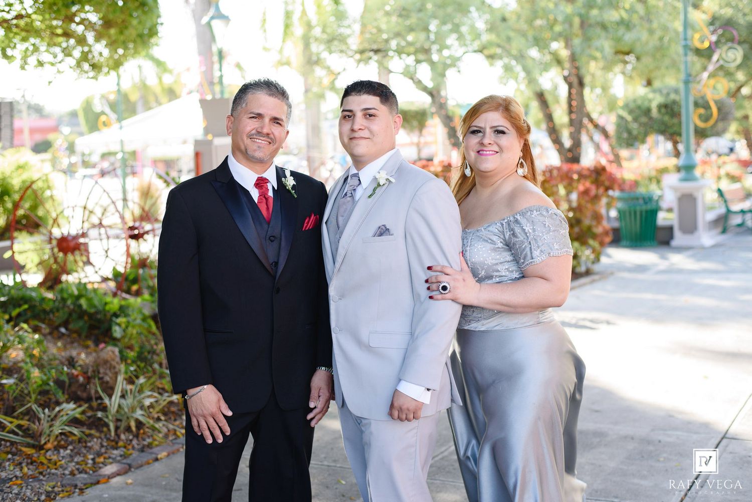 Family portrait with man in black tuxedo, young man in gray suit, and woman in silver gown outdoors.