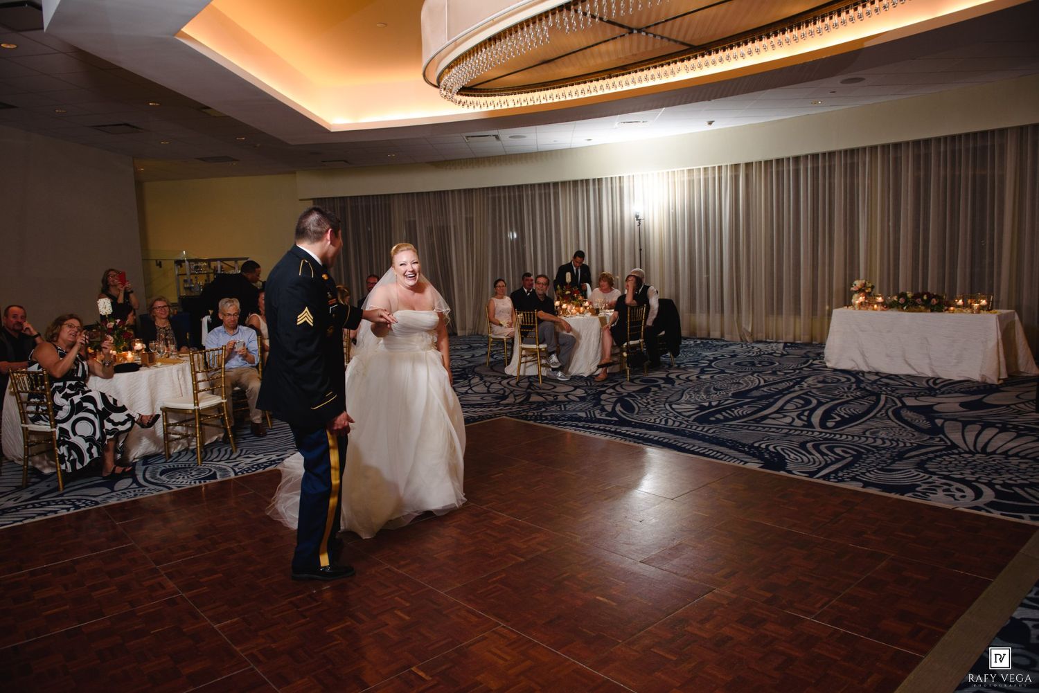 Bride and groom share their first dance at an elegant wedding reception in a ballroom with guests seated around.
