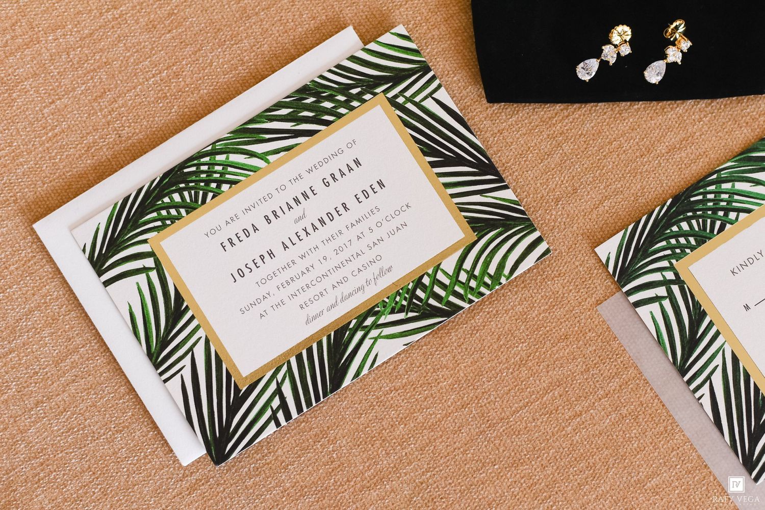 Tropical palm leaf wedding invitation with gold border, displayed alongside pearl earrings on a neutral background.