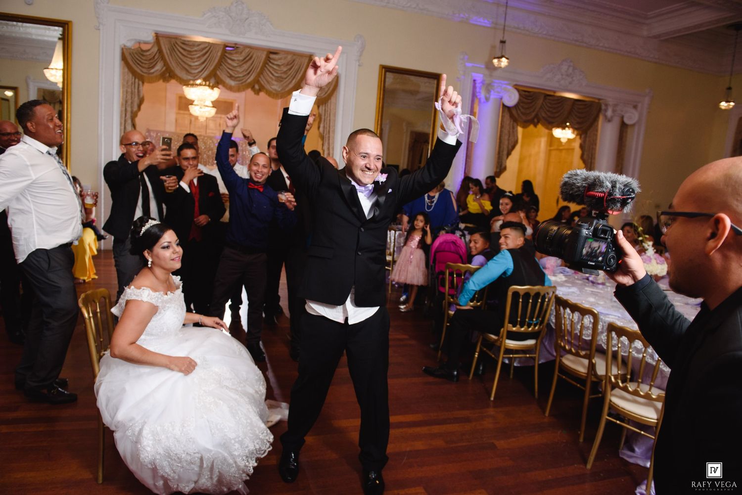 Groom celebrating with raised arms at wedding reception while bride sits in gown, guests and decorated ballroom in background.