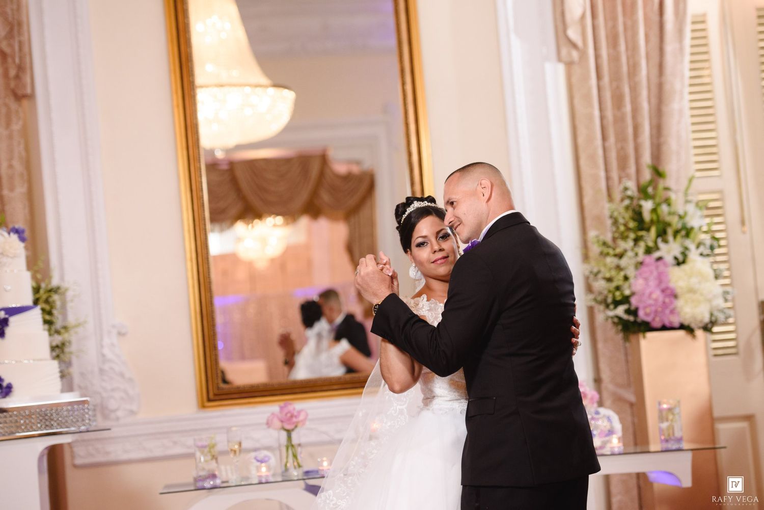 Bride and groom share their first dance in an elegant ballroom with a gold mirror and wedding cake in the background.
