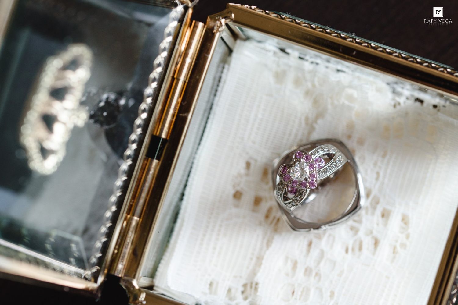 Silver ring with pink gemstone accents displayed in an open gold-trimmed jewelry box lined with white fabric.