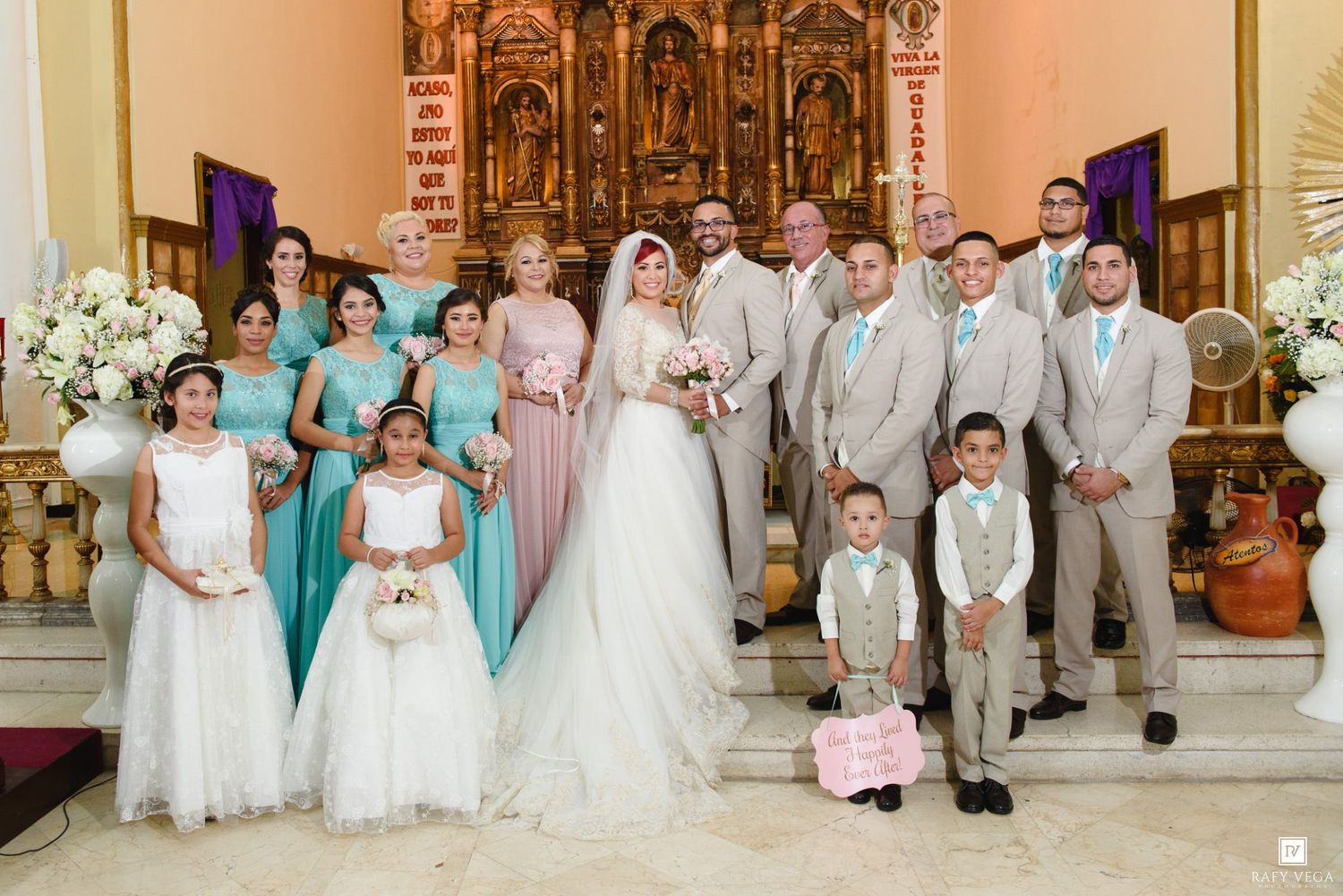 Wedding party poses in a church with bridesmaids in teal dresses, groomsmen in tan suits, and bride in white gown.