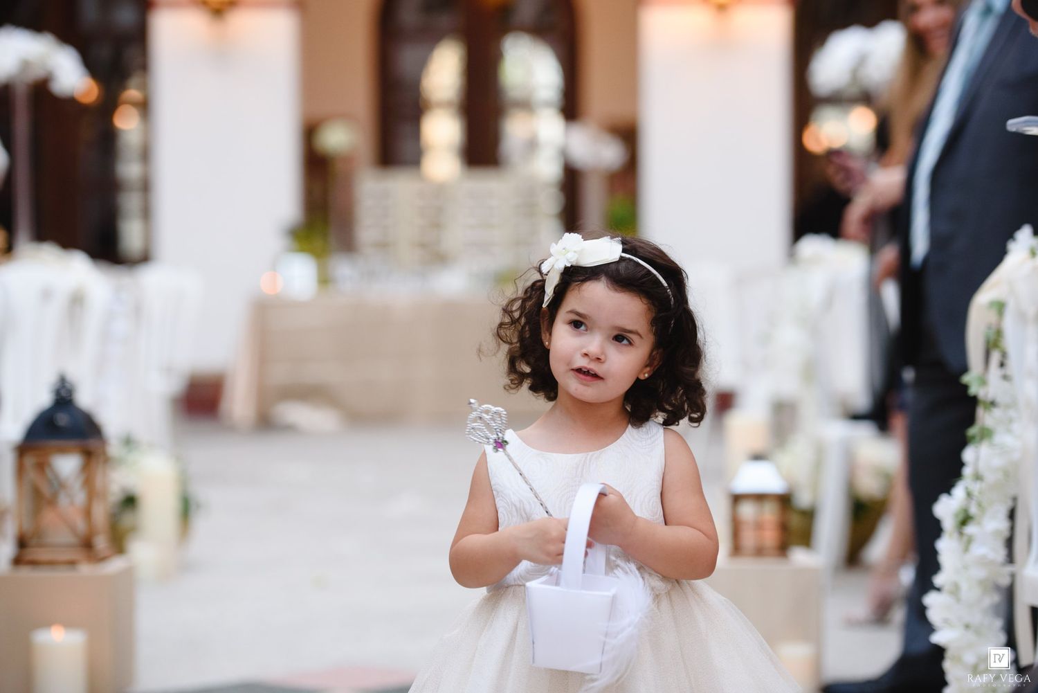 A young flower girl in a white dress holds a basket and wand at an elegant wedding ceremony.