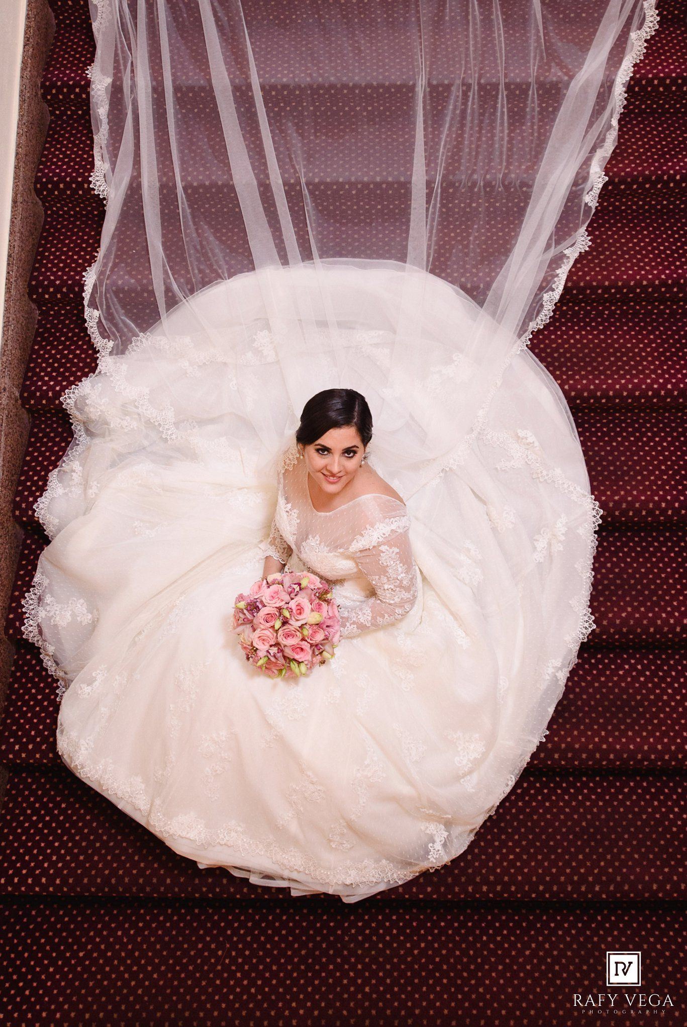 Aerial view of bride in white ball gown with lace veil holding pink bouquet on red carpeted stairs.