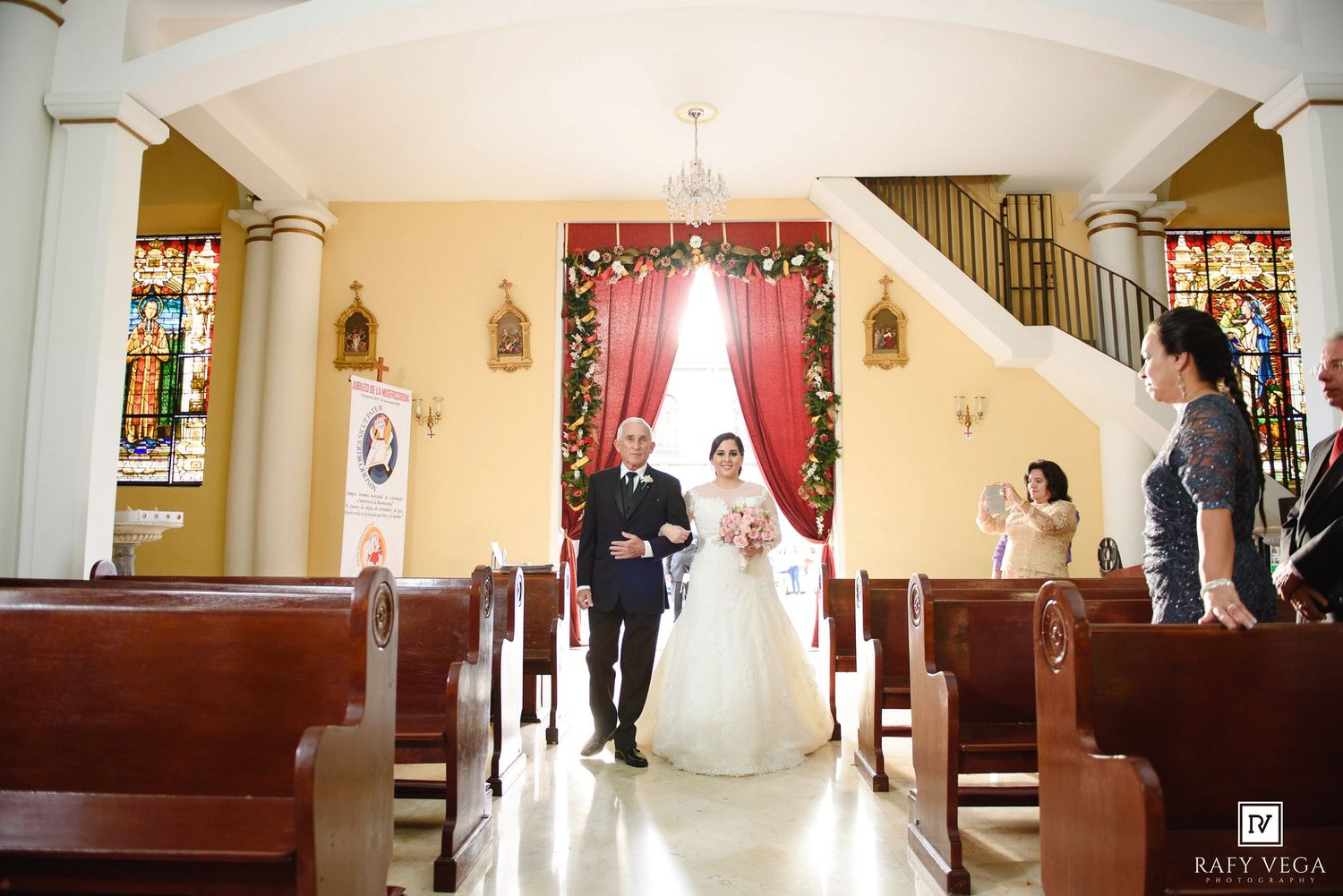 Bride in white gown walks down church aisle with escort during elegant wedding ceremony with floral decorations.