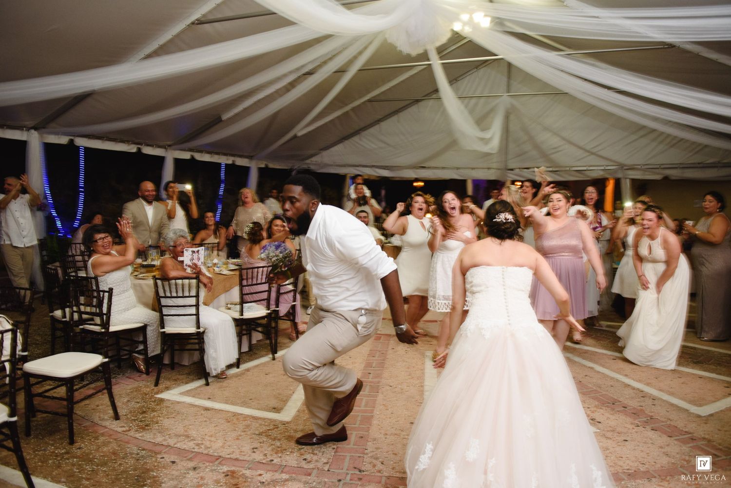Bride and groom dancing energetically at elegant wedding reception under draped white tent with guests watching.