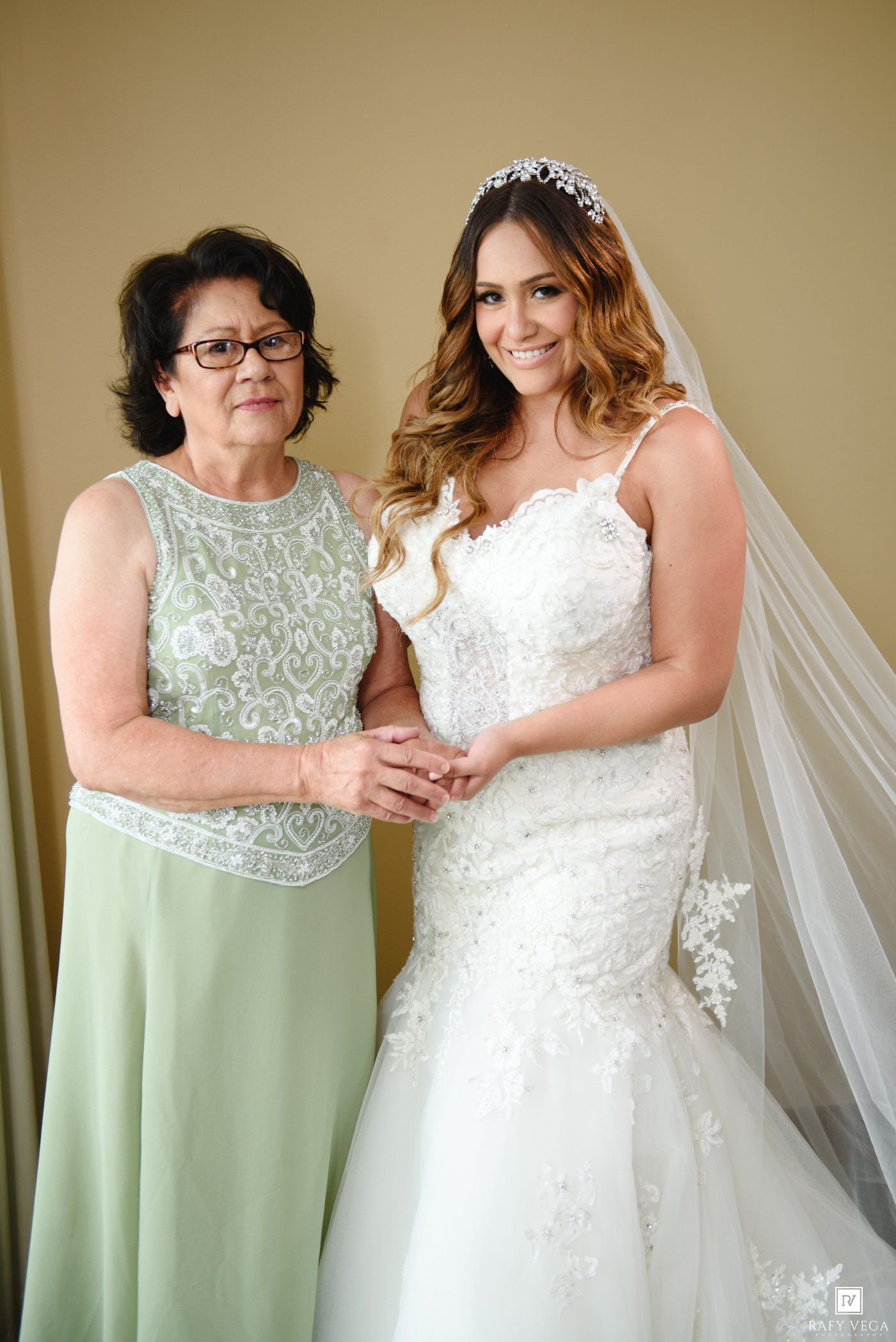 Bride in white lace mermaid gown and veil poses with older woman in sage green dress, holding hands.