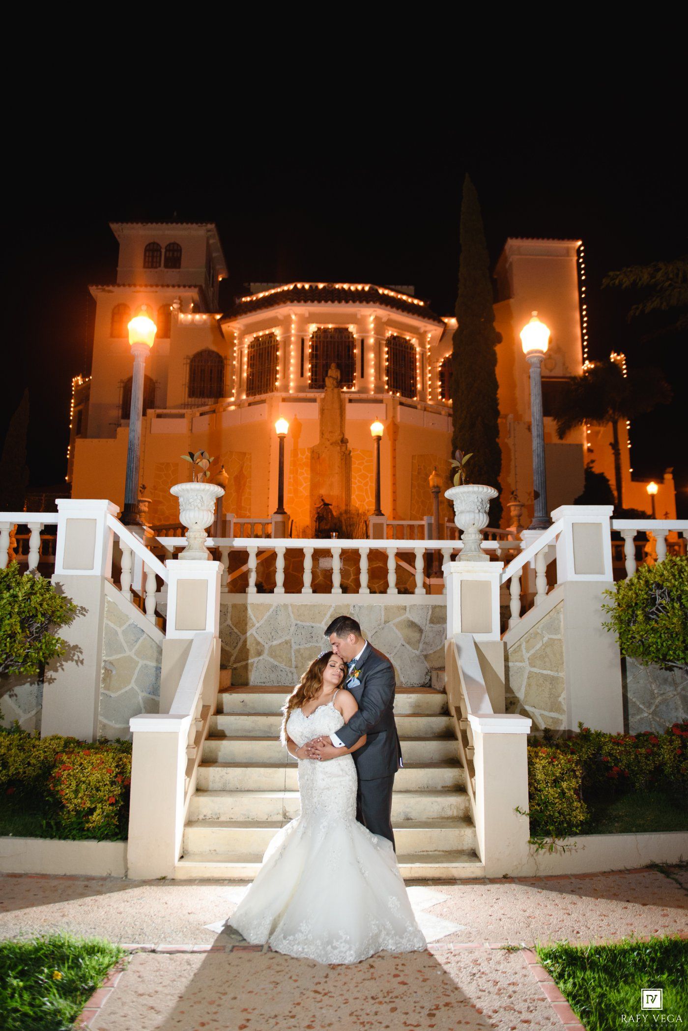 Bride and groom embrace at night on elegant staircase of illuminated wedding venue with warm golden lights.