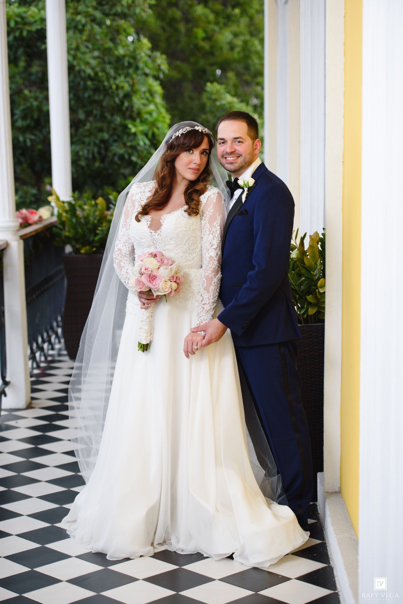 Bride and groom pose on a black and white checkered porch, bride in lace gown with veil holding pink bouquet.