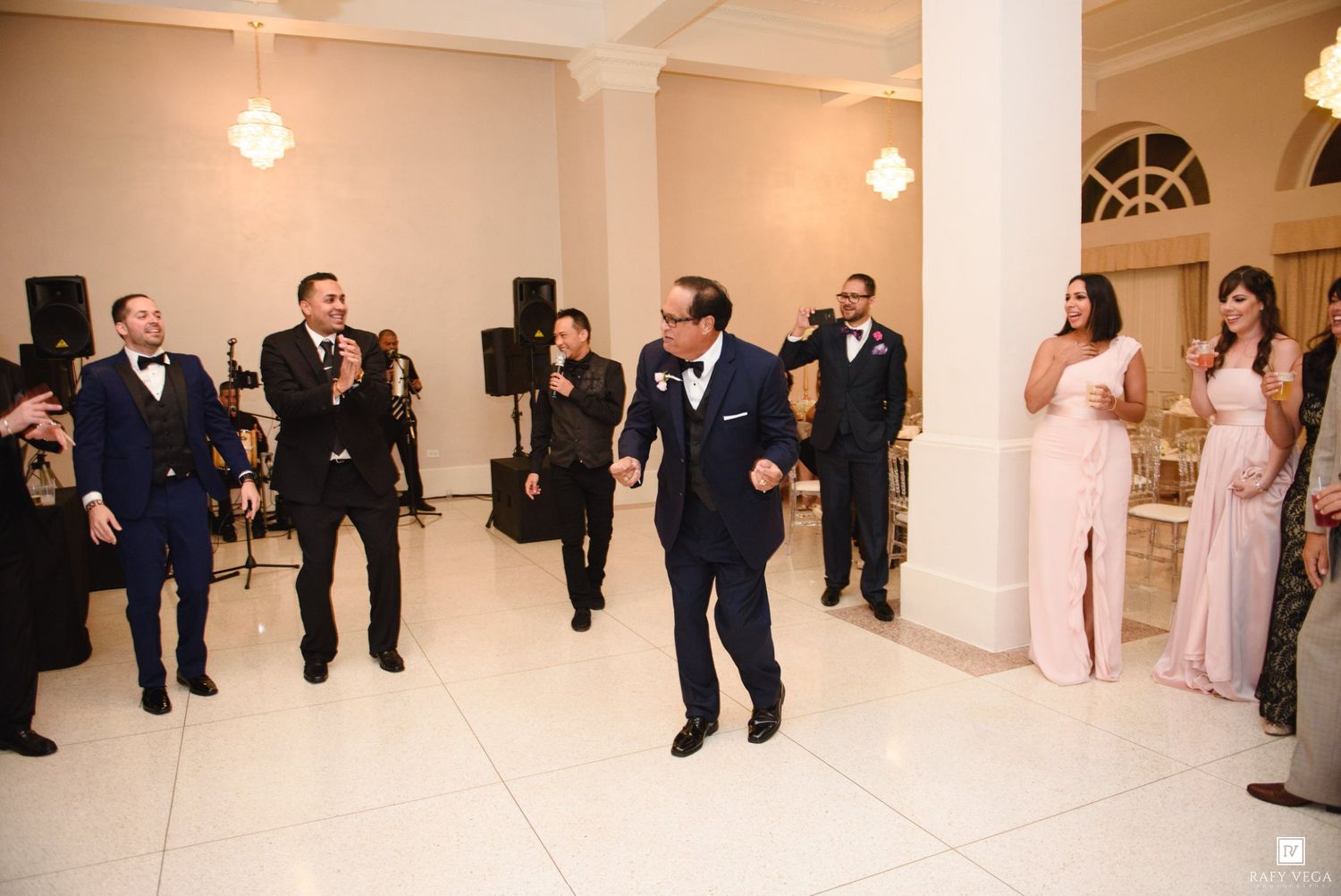 Groomsmen dancing at elegant wedding reception with bridesmaids in pink dresses watching and clapping.