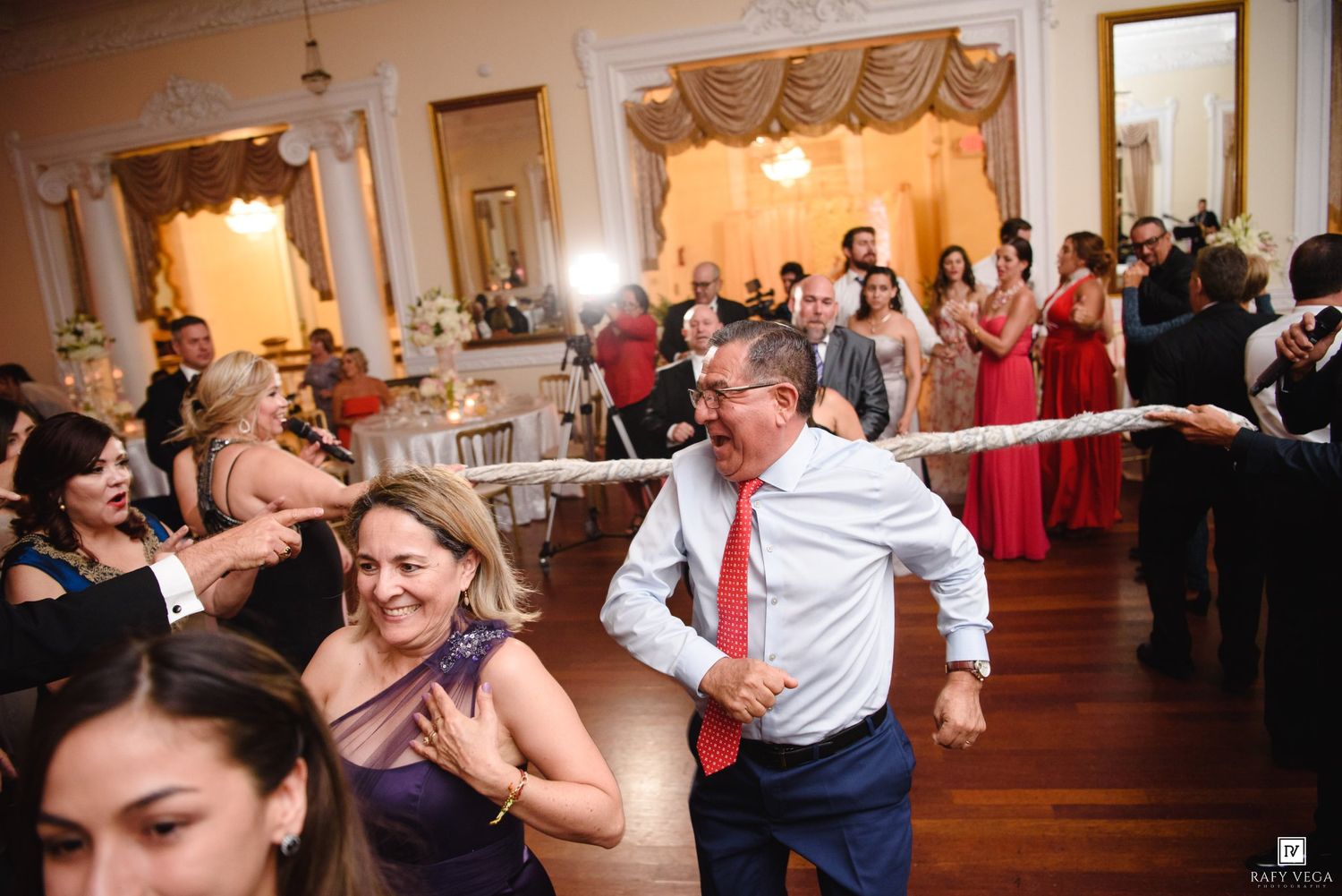 Guests laughing and dancing at an elegant wedding reception in a grand ballroom with chandeliers and formal decor.