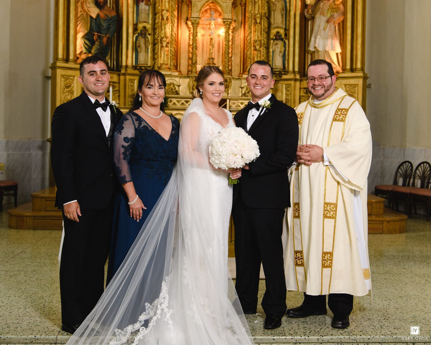 Wedding party poses with priest at ornate church altar, bride in white gown holding bouquet with family in formal attire.