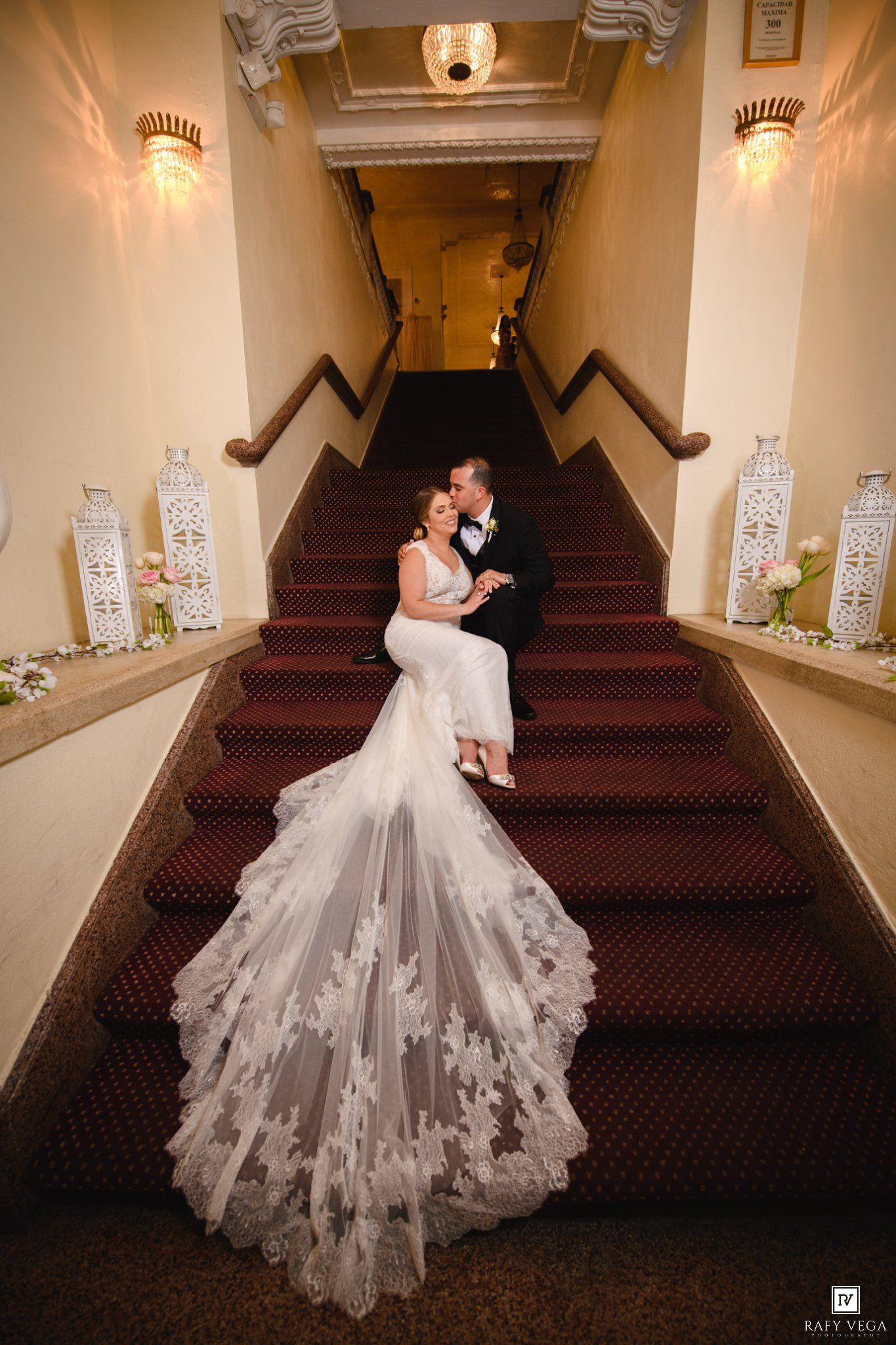 Wedding couple poses romantically on red-carpeted staircase, bride's lace cathedral veil cascading down the steps.