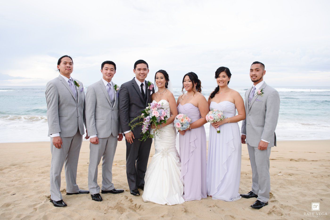 Wedding party poses on a sandy beach with groomsmen in gray suits and bridesmaids in lavender gowns.