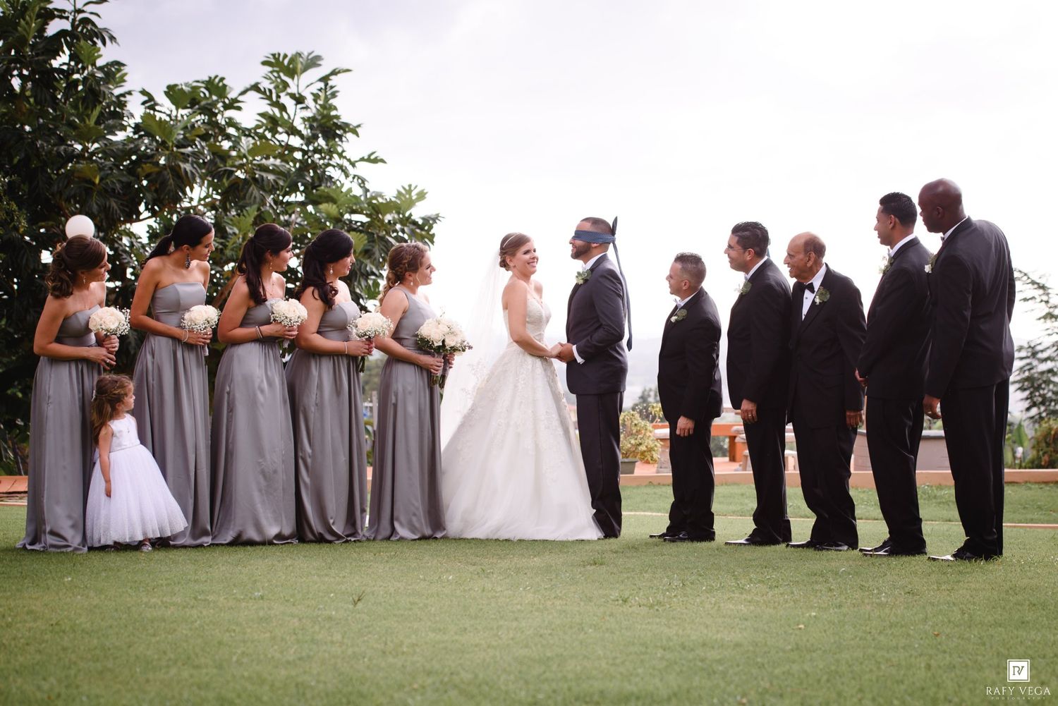 Wedding party with bridesmaids in gray dresses and groomsmen in black tuxedos flanking the bride and groom outdoors.