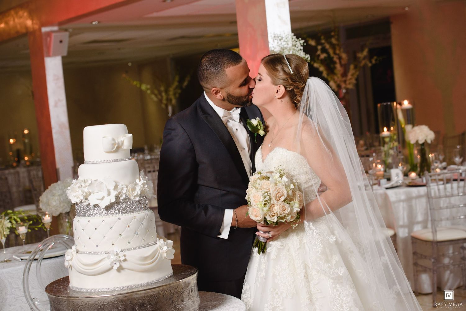 Newlywed couple sharing a kiss beside a white wedding cake in an elegantly decorated reception hall.