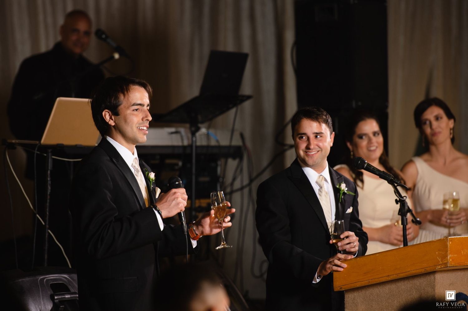 Man giving a toast speech at an elegant wedding reception while groom holds a drink at the podium.
