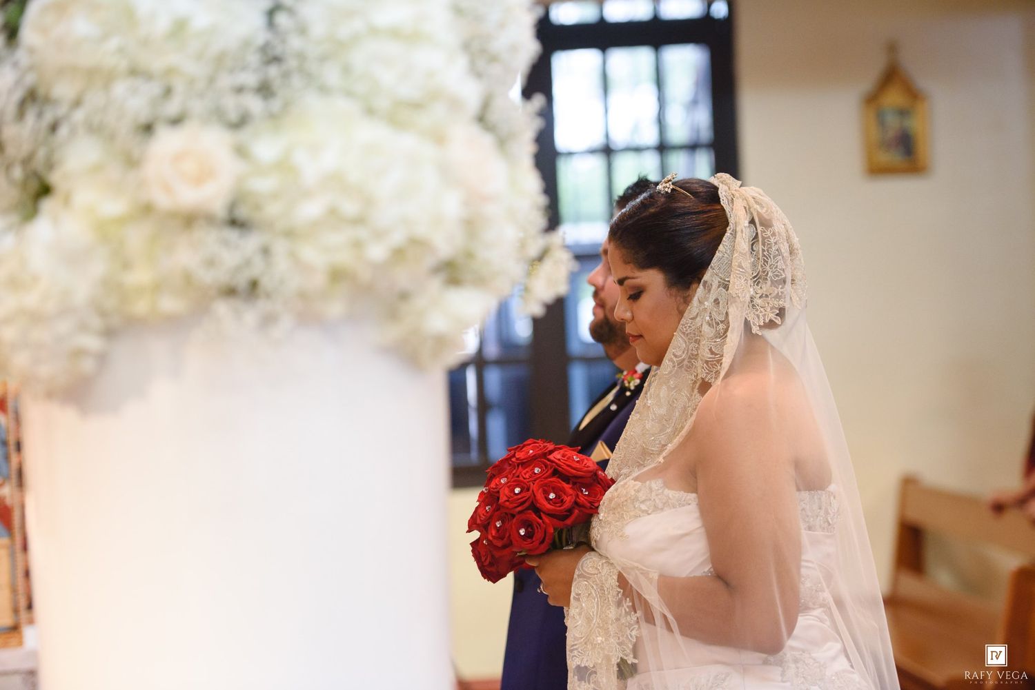 Bride in white gown and lace veil holds red rose bouquet at church altar with white floral arrangements.