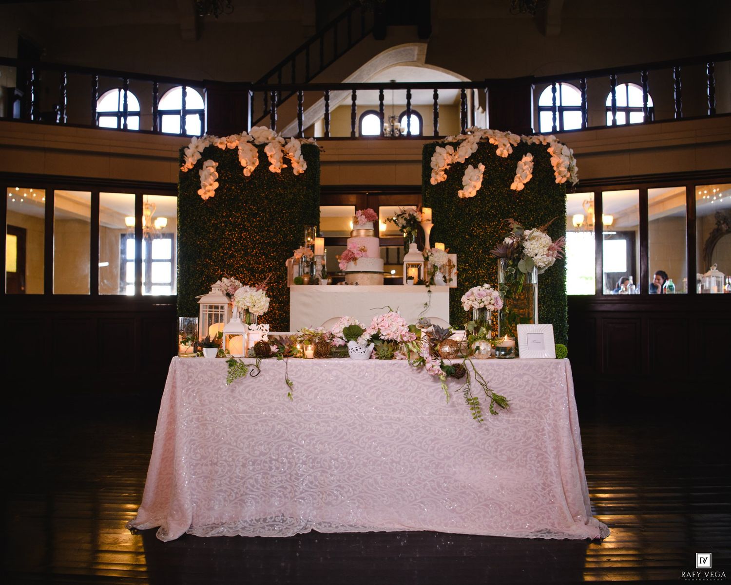 Elegant wedding sweetheart table with blush sequin linen, floral arch, candles, and cake display in a grand ballroom.