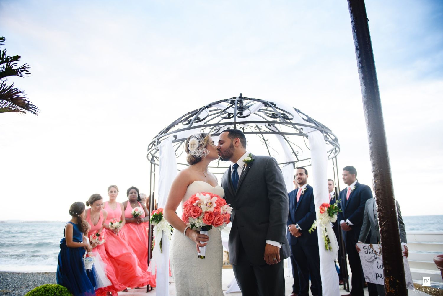 Bride and groom share a kiss at outdoor beach wedding ceremony under iron gazebo with bridesmaids in coral dresses.
