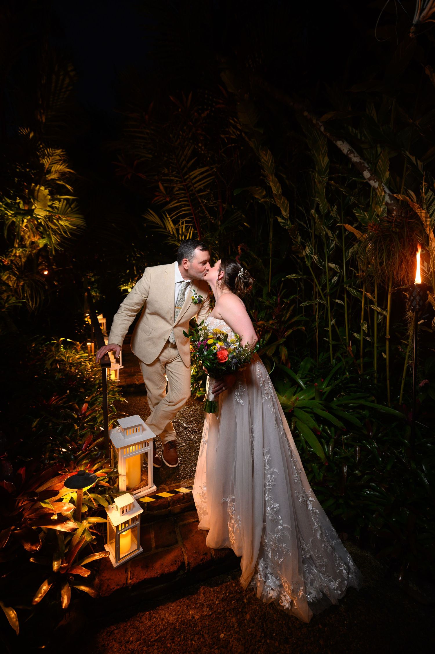 Bride and groom share a romantic kiss at night surrounded by tropical greenery and glowing lanterns.