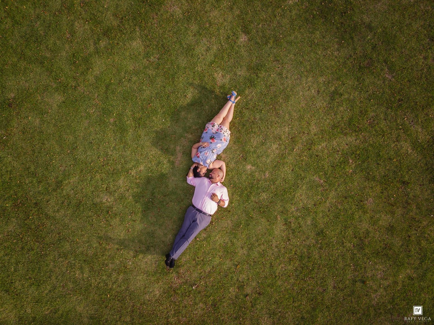 Aerial drone view of a couple lying on lush green grass, heads together in a romantic outdoor photo shoot.