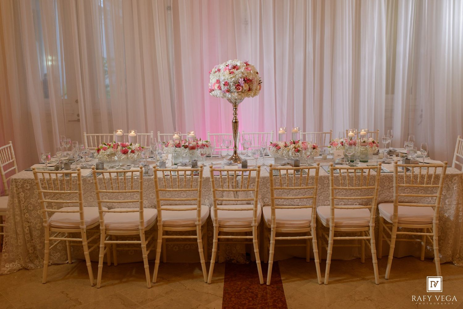 Luxury wedding reception table with gold chiavari chairs, tall floral centerpiece, and elegant place settings under white draping.