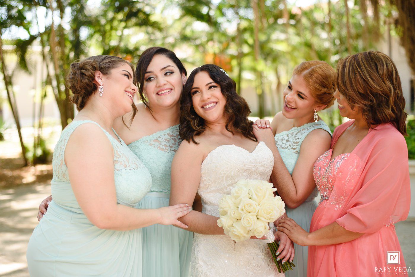 Bride in white gown holds bouquet surrounded by bridesmaids in mint green dresses and a woman in coral outdoors.
