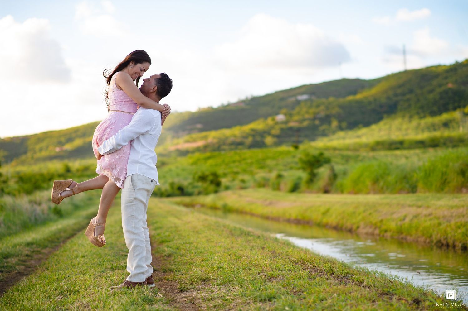 A man lifts a woman in a pink dress outdoors near a stream with lush green hills in the background.