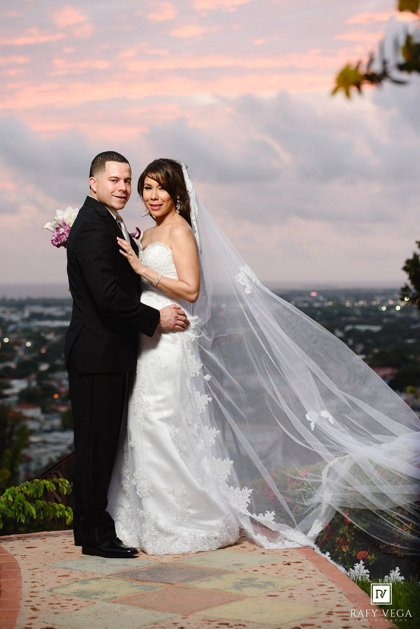 Bride and groom pose at sunset overlook, her lace veil flowing in the wind with city lights below.