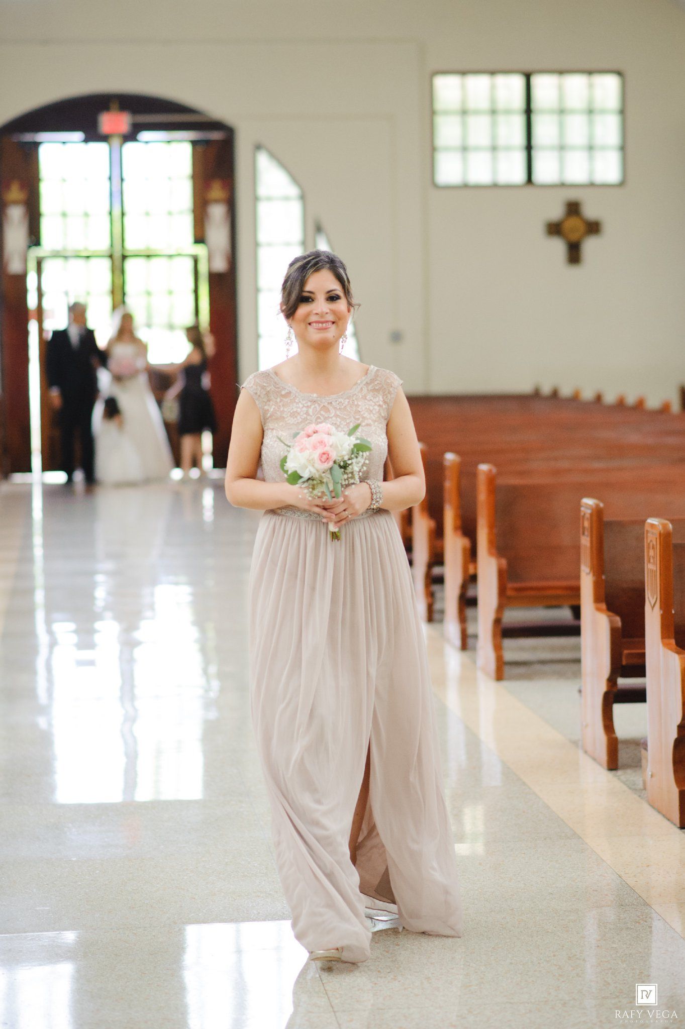 Bridesmaid in elegant taupe gown holding pink bouquet walks down church aisle during wedding ceremony.