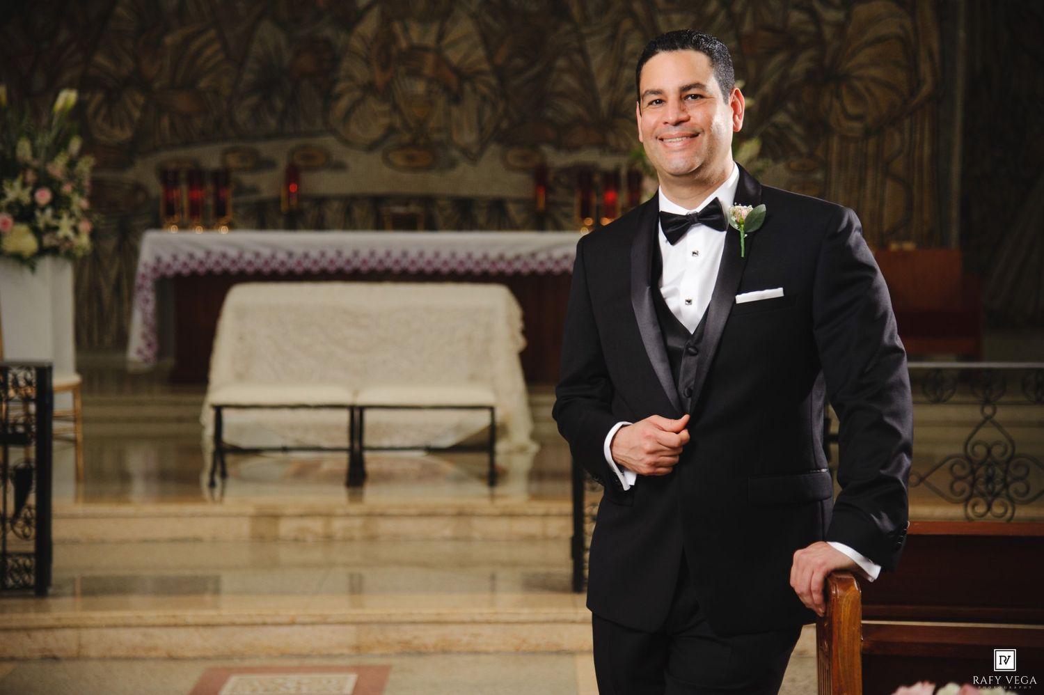 Handsome groom in black tuxedo with bow tie smiling inside ornate church with decorative altar backdrop.