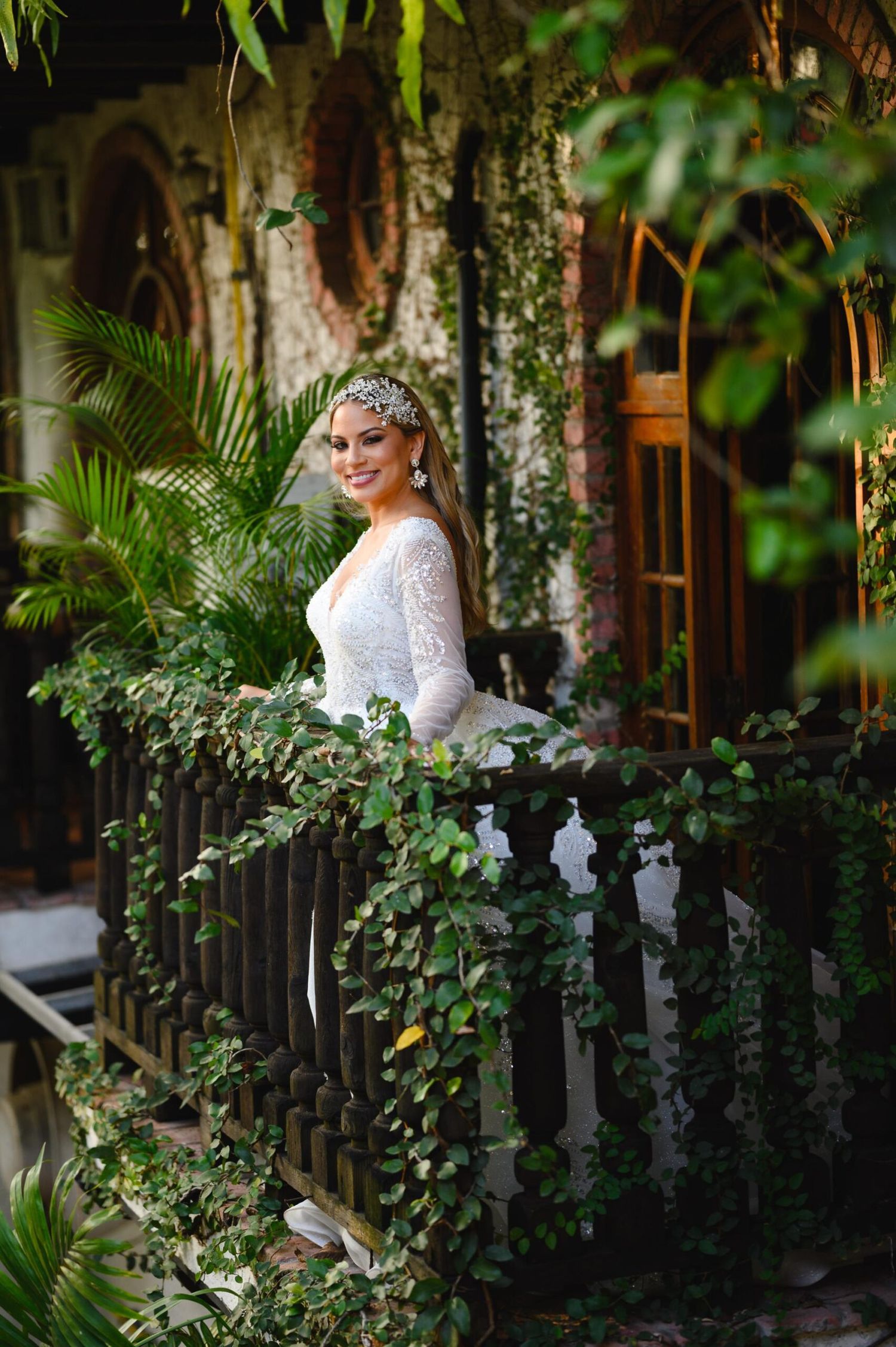 Bride in white lace gown and tiara smiles on ivy-covered balcony surrounded by lush tropical greenery.