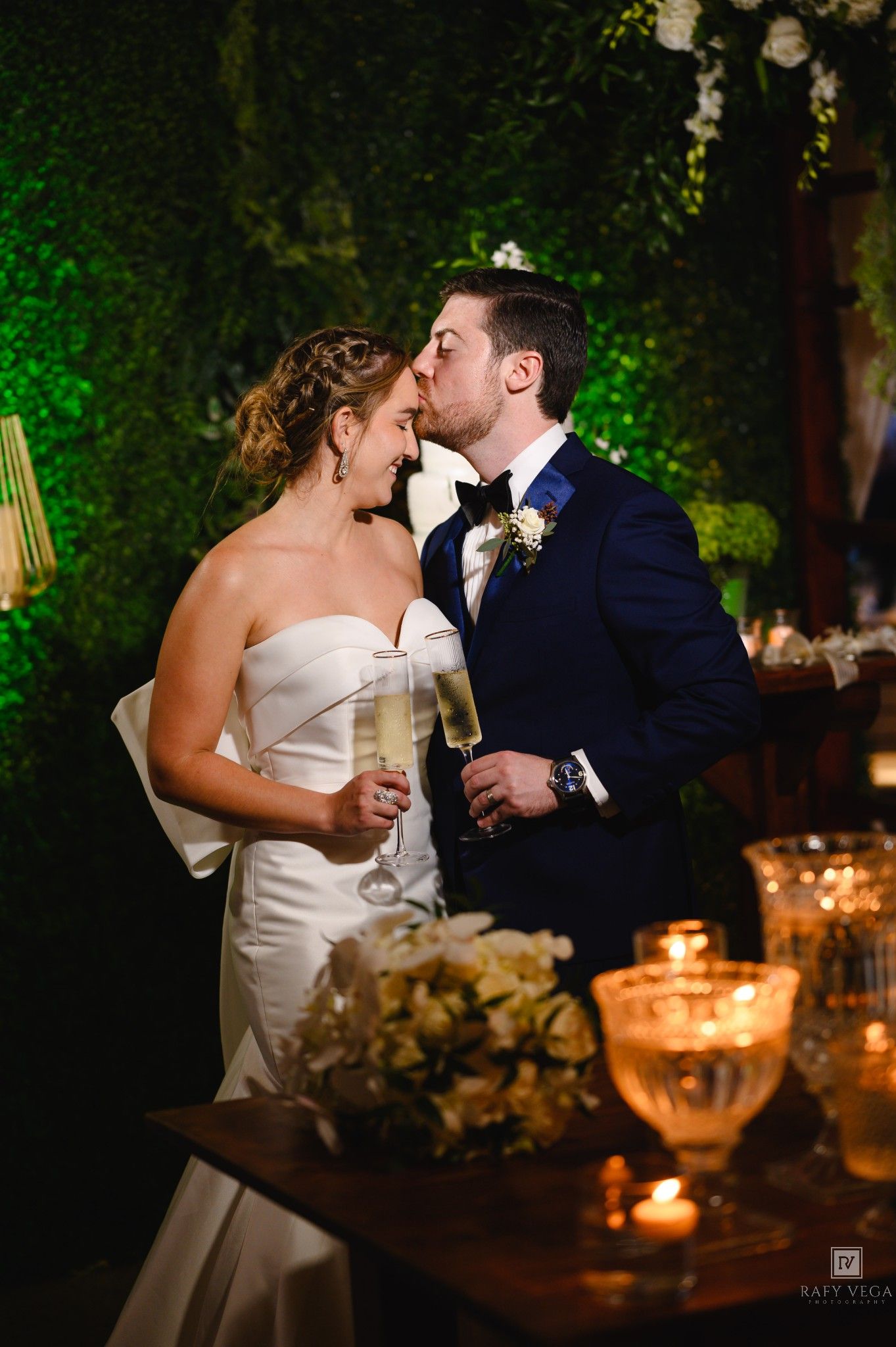 Bride and groom share a kiss while holding champagne glasses at their elegant candlelit wedding reception.