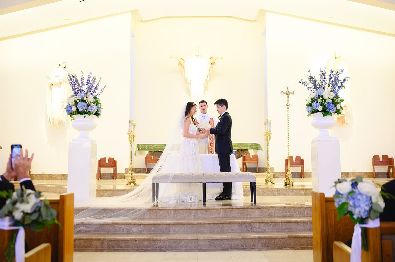 Bride and groom exchange vows at church altar adorned with white and blue floral arrangements during wedding ceremony.