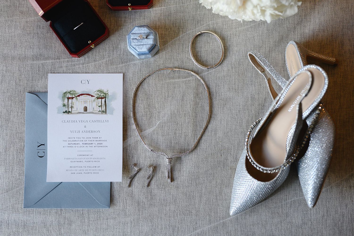 Elegant wedding flat lay featuring silver glitter heels, pearl necklace, bracelets, illustrated invitation, and jewelry boxes on gray linen.