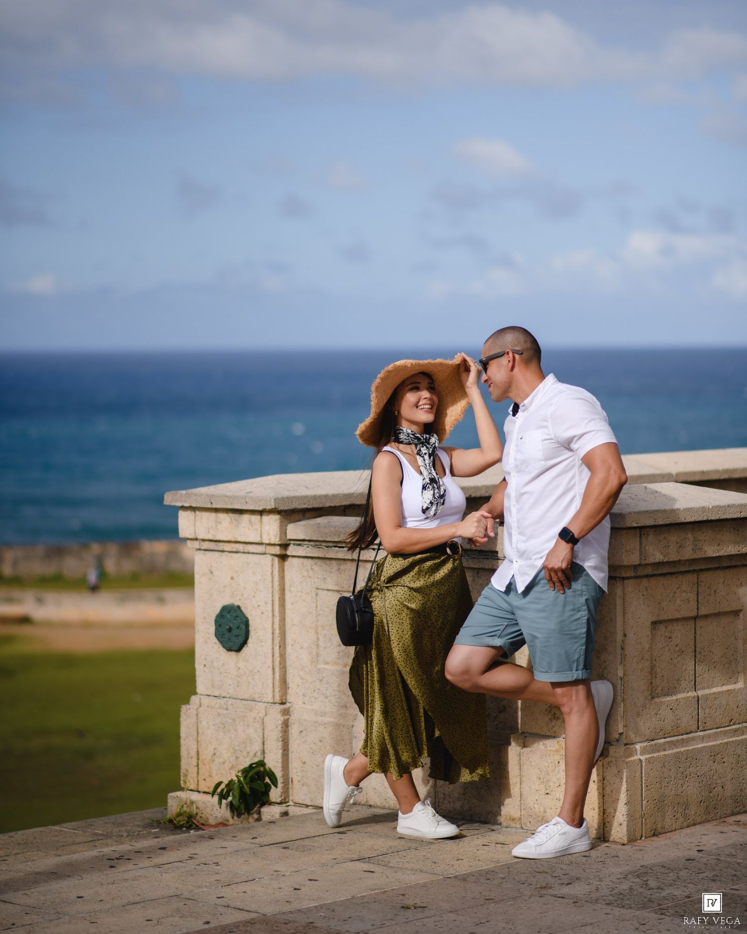 Couple posing by a stone wall overlooking the ocean on a sunny day, woman in hat and green skirt, man in white shirt.