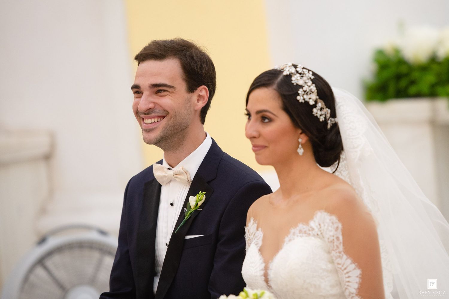 Smiling groom in tuxedo and bride in lace wedding dress with floral headpiece at their wedding ceremony.